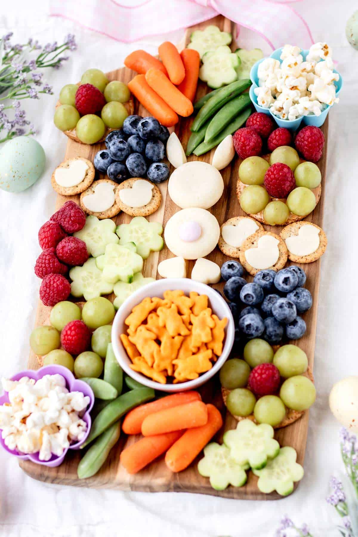 A wooden board with an Easter bunny made out of baby bel cheese, surrounded by various snacks like grapes, raspberries, carrots, and bunny-shaped crackers.