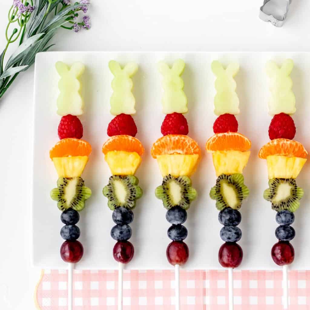A row of Easter fruit kabobs lined up on a white tray with a pink checkered napkin.